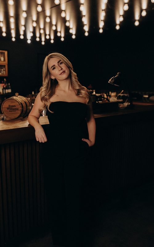 Laura Ritter lehnt elegant an einer Bartheke unter stimmungsvollem Licht, in einem schwarzen Outfit.